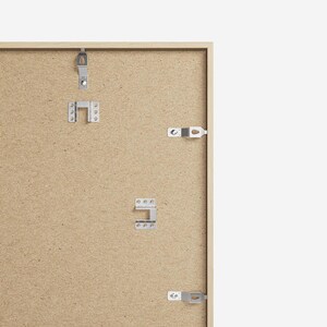 May include: The back of a light brown picture frame with metal hanging hardware attached. The frame is made of wood and has a textured surface.