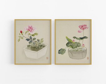 May include: Two framed watercolor paintings of flowers in pots. The painting on the left features a pink rose in a white pot. The painting on the right features a pink lotus flower in a gray pot.