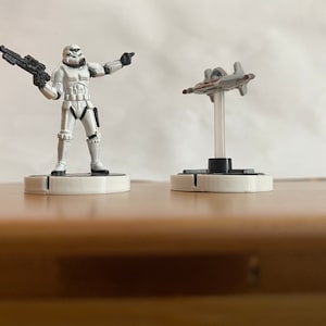 Puede incluir: Una figura de Stormtrooper blanca sosteniendo un rifle bl&aacute;ster negro se encuentra sobre una base blanca. Una figura de nave espacial gris y roja est&aacute; sobre una base blanca.