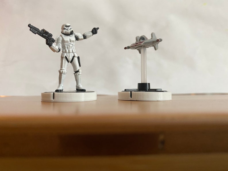 Puede incluir: Una figura de Stormtrooper blanca sosteniendo un rifle bl&aacute;ster negro se encuentra sobre una base blanca. Una figura de nave espacial gris y roja est&aacute; sobre una base blanca.