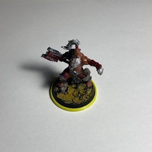 Puede incluir: Una figura miniatura pintada de un guerrero con un hacha roja y plateada, que lleva un taparrabos marr&oacute;n y una barba blanca. La figura est&aacute; de pie sobre una base marr&oacute;n y amarilla.