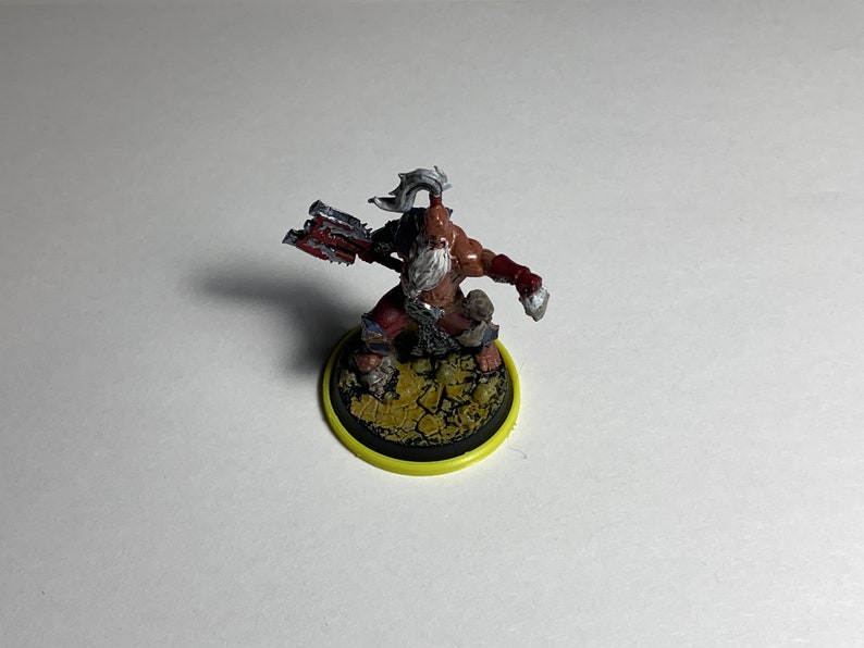 Puede incluir: Una figura miniatura pintada de un guerrero con un hacha roja y plateada, que lleva un taparrabos marr&oacute;n y una barba blanca. La figura est&aacute; de pie sobre una base marr&oacute;n y amarilla.