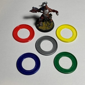 Pode incluir: Cinco anéis de plástico de diferentes cores, vermelho, amarelo, cinza, azul e verde. Os anéis são todos do mesmo tamanho e têm um furo no centro. Eles estão dispostos em círculo em torno de uma figura em miniatura.