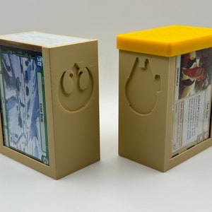 Puede incluir: Dos cajas de tarjetas impresas en 3D de color beige con un tema de Star Wars. Una caja tiene un símbolo de la Alianza Rebelde y la otra tiene un símbolo de la Estrella de la Muerte. La caja con el símbolo de la Estrella de la Muerte tiene una tapa amarilla.