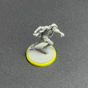 Puede incluir: Una figura en miniatura de pl&aacute;stico gris de una persona que lleva una chaqueta con capucha y corre. La figura est&aacute; de pie sobre una base gris con un anillo amarillo alrededor del borde.
