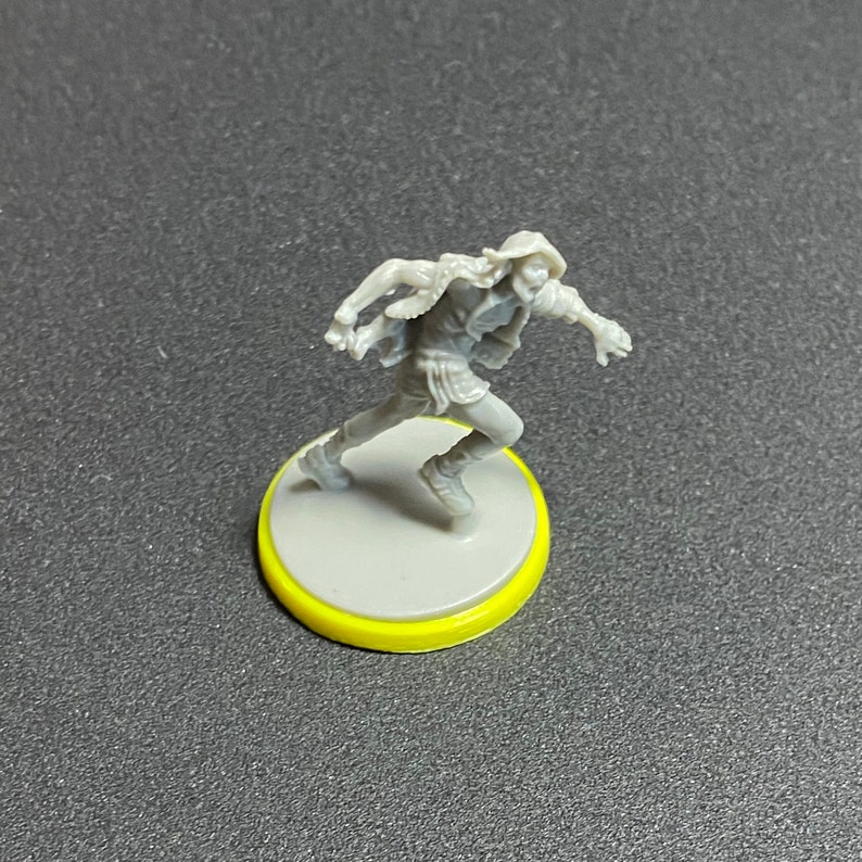Puede incluir: Una figura en miniatura de pl&aacute;stico gris de una persona que lleva una chaqueta con capucha y corre. La figura est&aacute; de pie sobre una base gris con un anillo amarillo alrededor del borde.