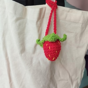 以下が含まれることがあります： 赤い手編みのイチゴのチャーム。緑の葉と黄色の種が付いており、赤いコードで吊り下げられています。 イチゴは白いトートバッグに取り付けられています。