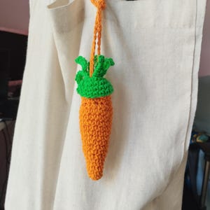 以下が含まれることがあります： 白いトートバッグに、緑の葉が付いた、かぎ針編みで作ったニンジンのキーホルダーがぶら下がっている。