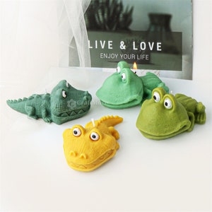 Könnte beinhalten: Vier neuartige Kerzen in Form von Cartoon-Alligatoren und Krokodilen. Die Kerzen sind in Grün- und Gelbtönen gehalten und haben weiße Dochte. Im Hintergrund befindet sich ein Schild mit der Aufschrift "LIVE & LOVE, ENJOY YOUR LIFE."