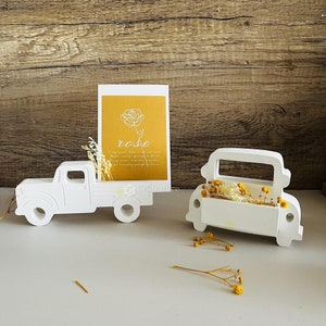 Könnte beinhalten: Zwei weiße Keramik-Lastwagen im Vintage-Design. Ein LKW trägt eine Karte mit dem Wort "Rose" und einen Trockenblumenstrauß. Der andere LKW trägt einen Trockenblumenstrauß.