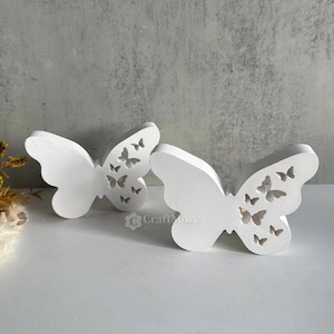 Könnte beinhalten: Zwei weiße Holz-Kerzenhalter in Form von Schmetterlingen mit ausgeschnittenen Schmetterlingsmotiven. Die Schmetterlinge schauen sich an.