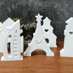 May include: Five white wooden cutout decorations of famous landmarks. The Eiffel Tower, the Burj Khalifa, a lighthouse, and two churches. Each cutout has a heart shape in the center.