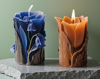 May include: Two decorative candles. One is blue with a floral design, the other is orange and lit. Both candles have a textured, bark-like base. They sit on a gray stone surface against a green background.