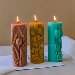May include: Three pillar candles with floral designs. The candles are in shades of brown, yellow, and teal. The candles are all lit and have a warm glow.