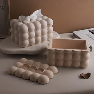 May include: A set of two rectangular beige tissue box covers featuring a raised bubble texture. One cover is assembled with white tissues protruding from the top, while the other is shown disassembled with its matching bubble-textured lid placed nearby.