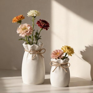 Op de afbeelding: Twee witte keramische vazen met een gerimpelde bovenkant en vastgebonden met een touwstrik. De vazen bevatten kleurrijke bloemen in tinten oranje, roze, geel en bordeaux. De vazen staan op een wit oppervlak.
