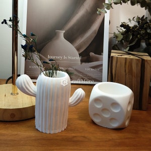 May include: Two white ceramic planters, one shaped like a cactus and the other like a dice. The cactus planter has a textured surface and a small opening at the top. The dice planter has six sides with raised dots. Both planters are on a wooden surface.