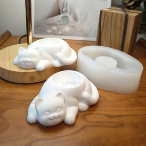 Può includere: Due stampi in silicone bianco per realizzare oggetti in resina. Uno stampo ha la forma di un gatto sdraiato con un'incisione rotonda sulla schiena. L'altro stampo è una semplice forma ovale. Gli stampi sono su una superficie di legno.