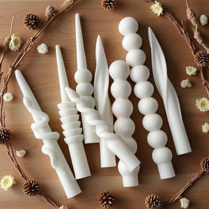 Può includere: Un assortimento di candele decorative bianche in varie forme, tra cui design attorcigliati, impilati e lisci. Le candele sono disposte su una superficie di legno, circondate da una ghirlanda di rametti, pigne e piccoli fiori bianchi. L'estetica generale è minimalista ed elegante.