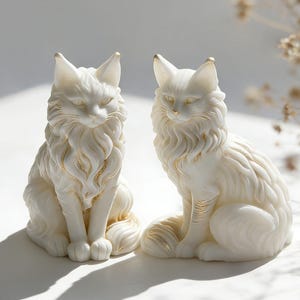 May include: Two white cat figurines with gold accents. The cats are in a seated position with detailed fur and closed eyes. The gold highlights the ears, chest, and tail. The figurines are on a white surface.