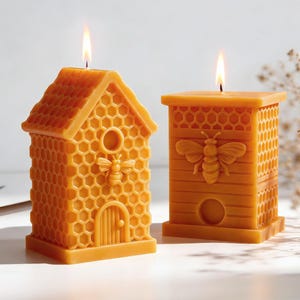 Könnte beinhalten: Zwei brennende, gelbe Bienenwachskerzen in Form eines Bienenstocks und eines Bienenkorbs. Die Bienenstockkerze hat eine Tür und eine Biene, während die Bienenkorbkerze eine Biene zeigt. Beide Kerzen haben ein Wabenmuster.