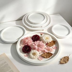 Peut inclure: Une collection de plateaux circulaires blancs à bords surélevés, dont un grand plateau rempli d'un arrangement de chrysanthèmes roses, bordeaux et blancs, accompagnés de plusieurs feuilles brunes séchées et de pétales éparpillés.