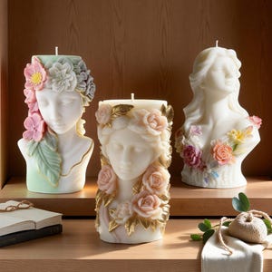 May include: Three decorative candles shaped like busts of women, adorned with floral designs. The candles feature a variety of colors, including white, pink, and green, and are arranged on a wooden shelf. The candles are approximately 6 inches tall.