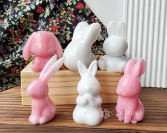May include: Six ceramic bunny figurines, three white and three pink, arranged on a wooden surface. The bunnies are in various poses, including sitting, standing, and holding their paws together.