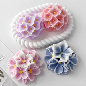 May include: A collection of floral-shaped soaps in shades of pink, purple, and blue. The soaps are arranged on a white, rope-edged tray and a white surface. Each soap features detailed petal designs with yellow centers.