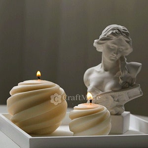 Può includere: Due candele bianche a spirale con una fiamma in cima, appoggiate su un vassoio bianco con una statua in gesso bianco di una donna con una corona di foglie sulla testa.