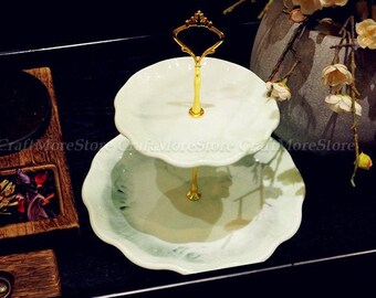 May include: Two-tiered white ceramic cake stand with a gold metal stand and a scalloped edge. The stand is on a black surface.
