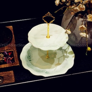 May include: Two-tiered white ceramic cake stand with a gold metal stand and a scalloped edge. The stand is on a black surface.