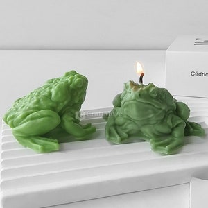 Könnte beinhalten: Zwei grüne Kerzen in Froschform, eine mit einer brennenden Docht, auf einer weißen Oberfläche. Die Kerzen sind detailliert und realistisch, mit strukturierter Haut und hervorstehenden Augen. Der Hintergrund zeigt eine weiße Schachtel mit dem Text "Cédric Viollet".