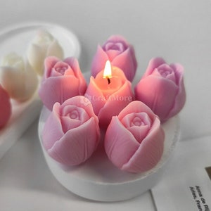 Op de afbeelding: Een set roze en witte roosvormige kaarsen op een wit bord. De kaarsen zijn aangestoken en geven een warm licht.