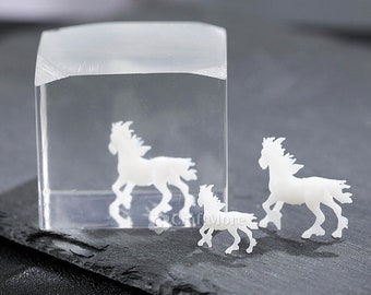 Op de afbeelding: Drie witte paardenfiguurtjes van hars, één in een doorzichtige kubus en twee op een zwart leisteen oppervlak. De paarden staan in een rennende houding.