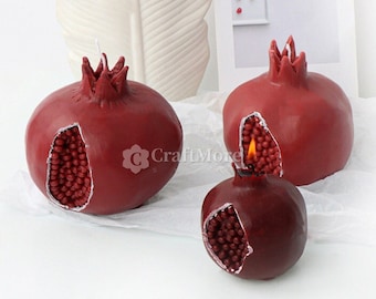 May include: Three red pomegranate shaped candles with a cut-out design revealing the seeds inside. The candles are on a white surface.