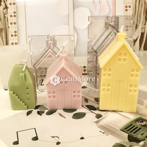 May include: Three small house-shaped candles in green, pink, and yellow. The candles are made in clear plastic moulds. The moulds are visible in the background.