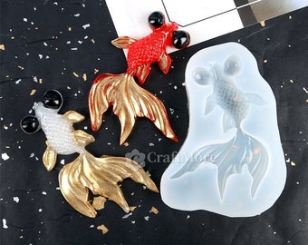 May include: Three gold and red resin goldfish figurines with black eyes. The figurines are sitting on a black surface next to a clear silicone mold of a goldfish.