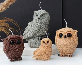 May include: Four owl-shaped candles in varying shades of brown and gray. The candles have detailed feather textures and large, dark eyes. Each candle has a white wick extending from the top. The candles are arranged on a white surface against a dark background.