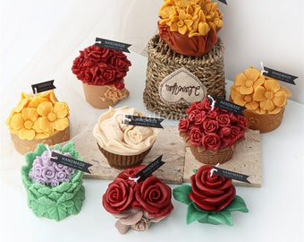 May include: Assortment of handmade flower candles in various colors and styles. The candles are arranged on a white surface with a brown wicker basket in the center. Each candle has a small black tag with the word "Handmade" written on it.