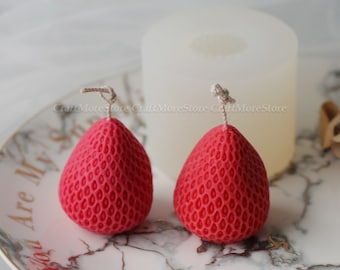 May include: Two red strawberry-shaped candles with a textured surface. The candles are on a white and gray marble-patterned plate.
