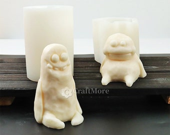 May include: A light-colored, handmade soap shaped like a cartoon monster, sitting next to a cylindrical mold and a monster-shaped mold. The soap has a smiling face and is sitting with its legs out. The background is a dark wooden surface.