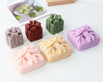 May include: Six decorative candles in the shape of wrapped gifts. The candles are in various colors, including purple, pink, green, brown, and beige. The candles are all tied with a ribbon and have a small flower on top.
