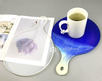 May include: Two resin serving boards, one clear and one blue. The clear board is shaped like a paddle with a handle and a hole. The blue board has a white handle and a white and blue design. A white mug with tea sits on the blue board.