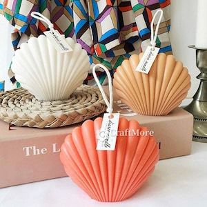 Può includere: Tre candele decorative a forma di conchiglia in bianco, arancione e rosa. Ogni candela ha un piccolo cartellino attaccato con il testo "shore candle".