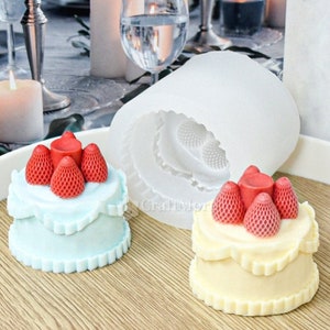 Op de afbeelding: Twee kleine taarten versierd met rode aardbeien en witte frosting. De taarten bevinden zich in een witte siliconen mal. De mal is gevormd als een taart met een gegolfde rand.
