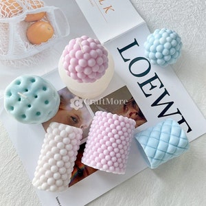 Peut inclure: Six moules à bougies différents en silicone. Les moules sont en forme de sphère, de cylindre et de rectangle. Les moules sont roses, bleus et blancs. Les moules sont sur une surface blanche avec le mot "LOEWE" imprimé dessus.