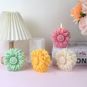 May include: Four decorative flower-shaped candles in different colors: green, yellow, white, and pink. The candles are arranged on a table with a book and a lamp in the background.