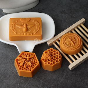 May include: Four honey-coloured soap bars with bee and honeycomb designs. One bar reads "QUEEN" in all caps. The soaps are on a white dish and a wooden soap dish.