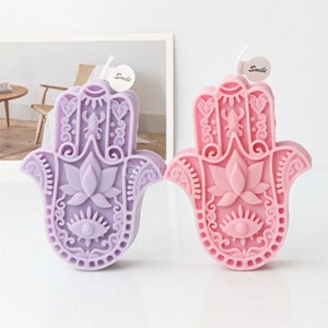Könnte beinhalten: Zwei Hamsa-Handkerzen, eine lila und eine rosa, mit aufwendigen Blumendesigns und einem bösen Auge in der Mitte. Jede Kerze hat ein "Smile"-Etikett.